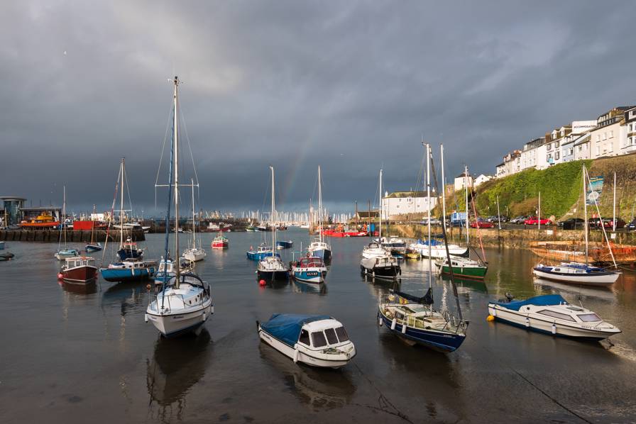 Boats floating in Brixham Harbour with a rainbow in a grey, cloudy sky.